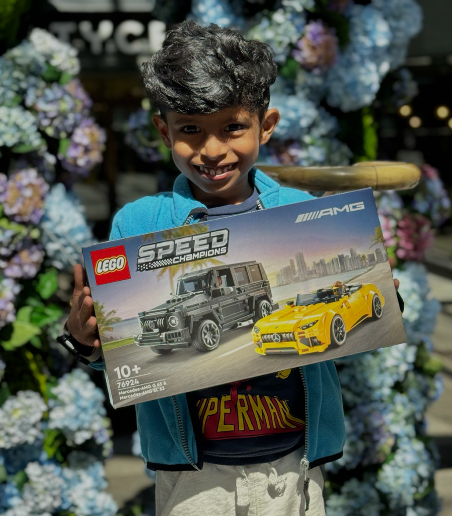 A child smiling at the camera, holding a Lego box with two cars on the cover.