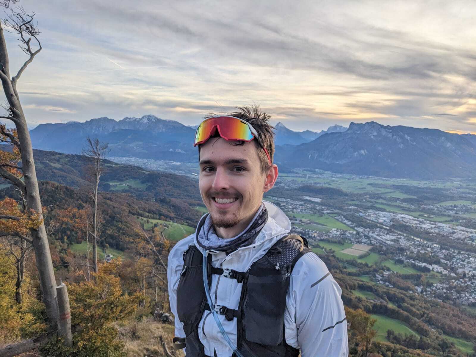 A person smiling towards the camera with a mountain landscape behind them.