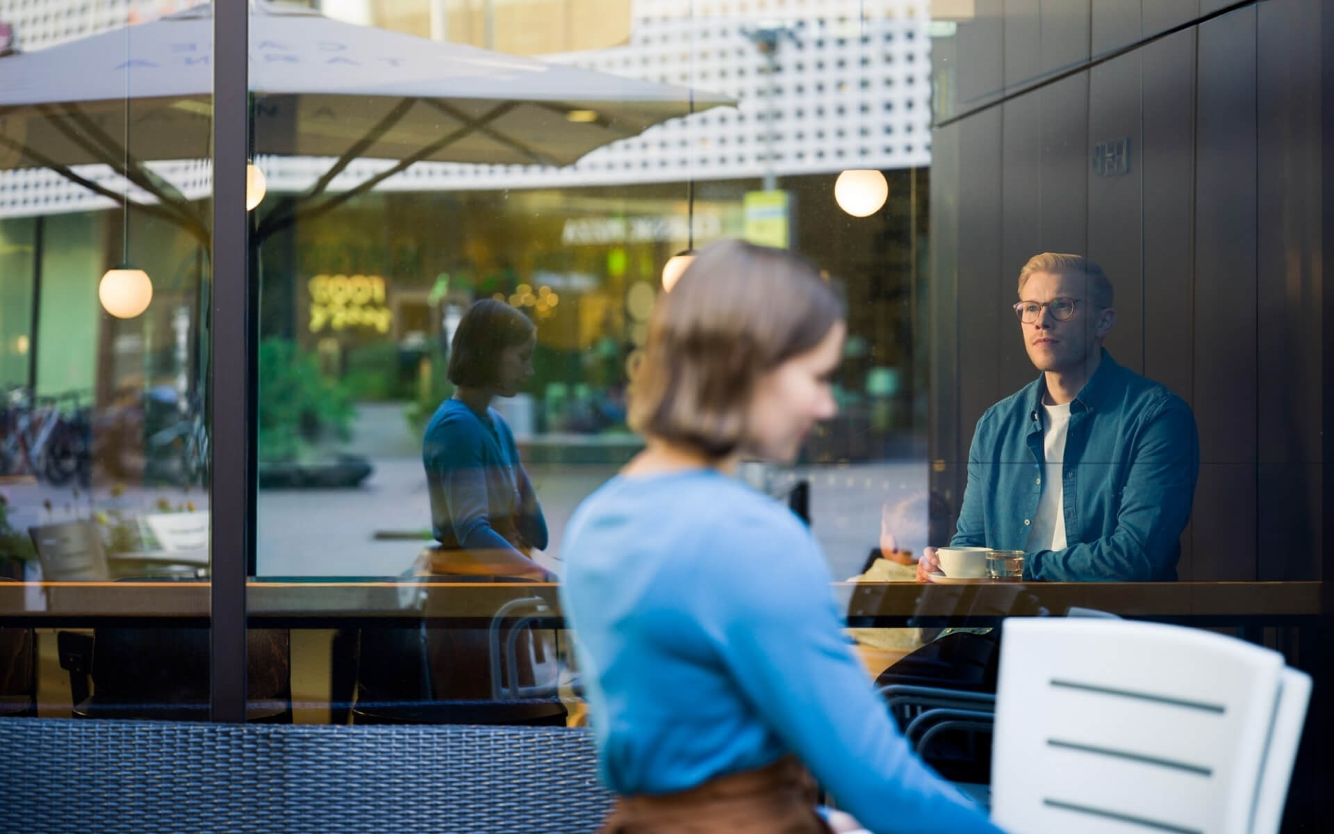 Waiter carring chairs outside while a man looks ahead behind the cafe window