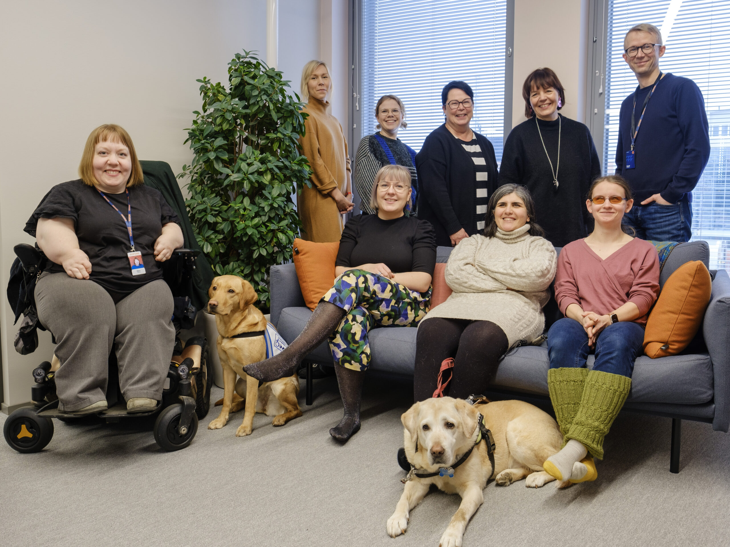 Three people sitting on a sofa. Five people are standing behind them and next to them is one person in a wheelchair. There are two guide dogs in front of the crew.
