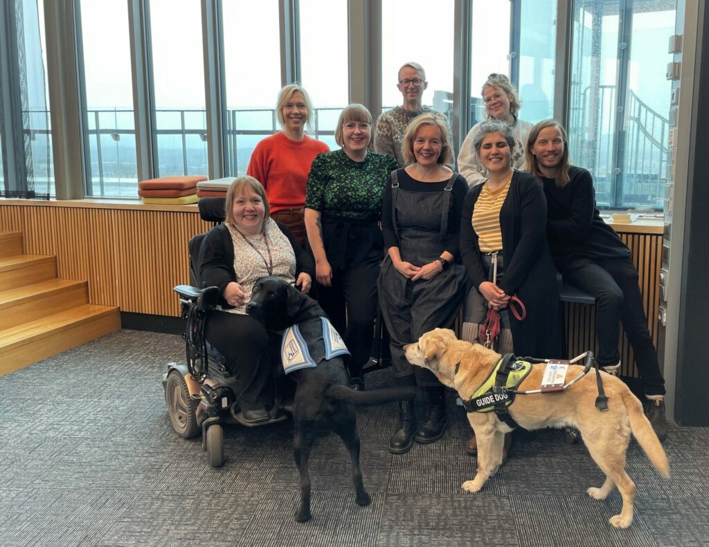 8 people smile for the camera in front of the window. One of the people uses a wheelchair, one has a white cane in her hand. In the foreground is one guide dog and one assistance dog. Three of the people have glasses.