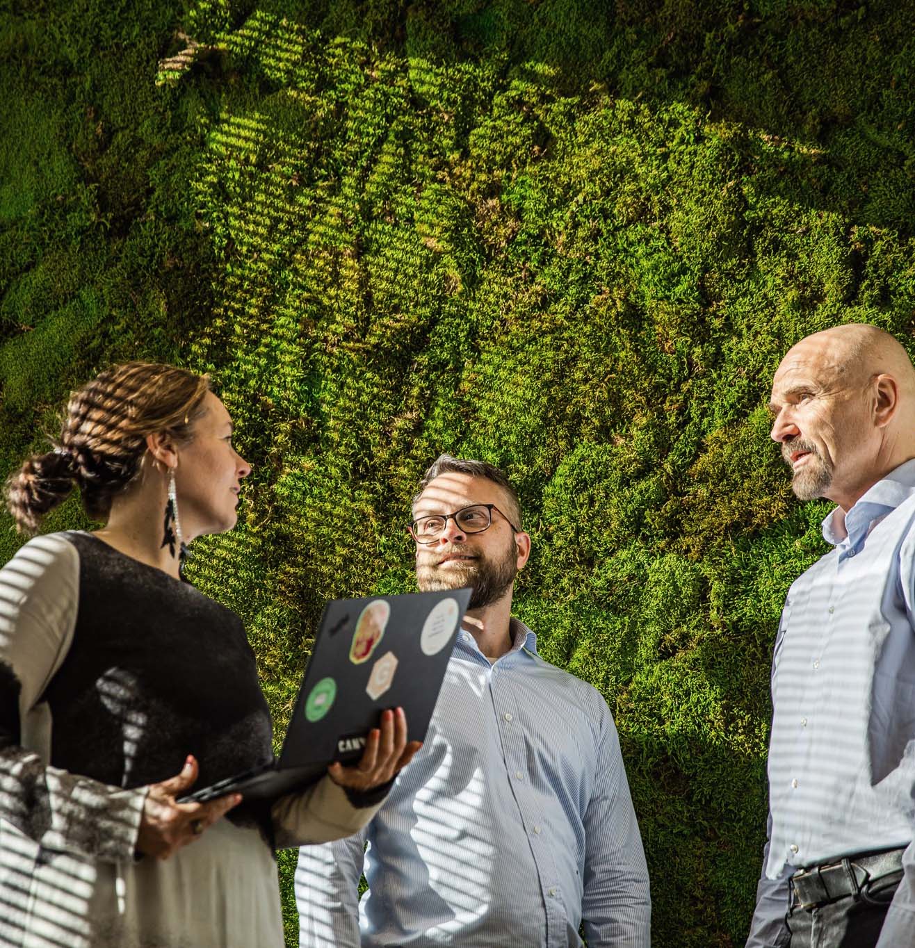 a woman and two men in front of a greenwall discussing